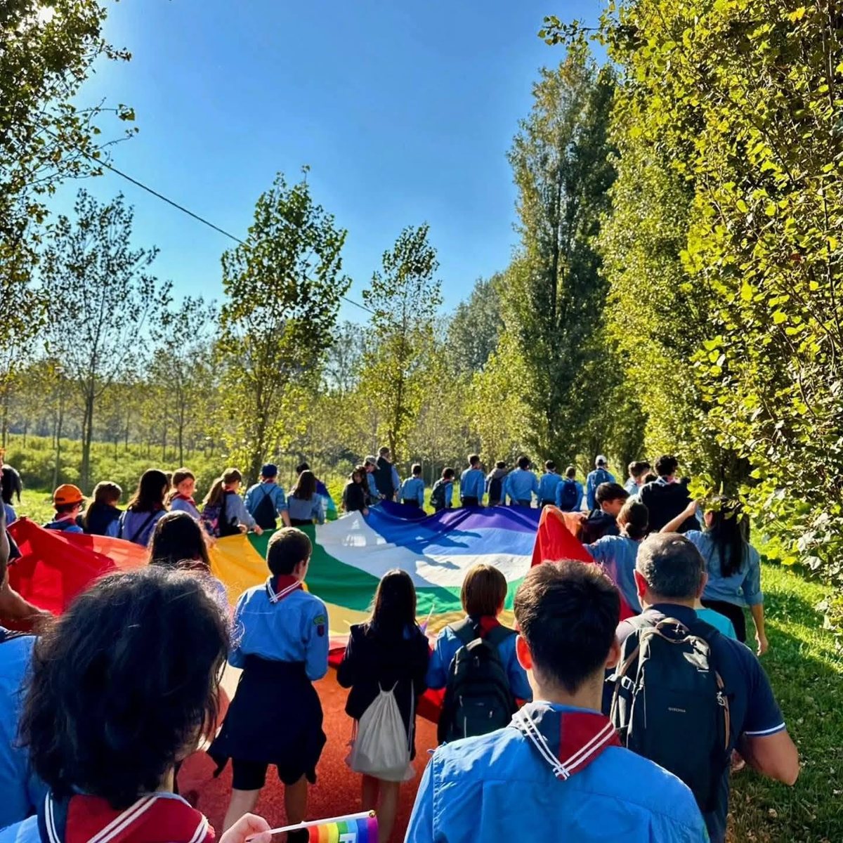 Bambini, scout e adulti camminano insieme sostenendo un grande telo arcobaleno